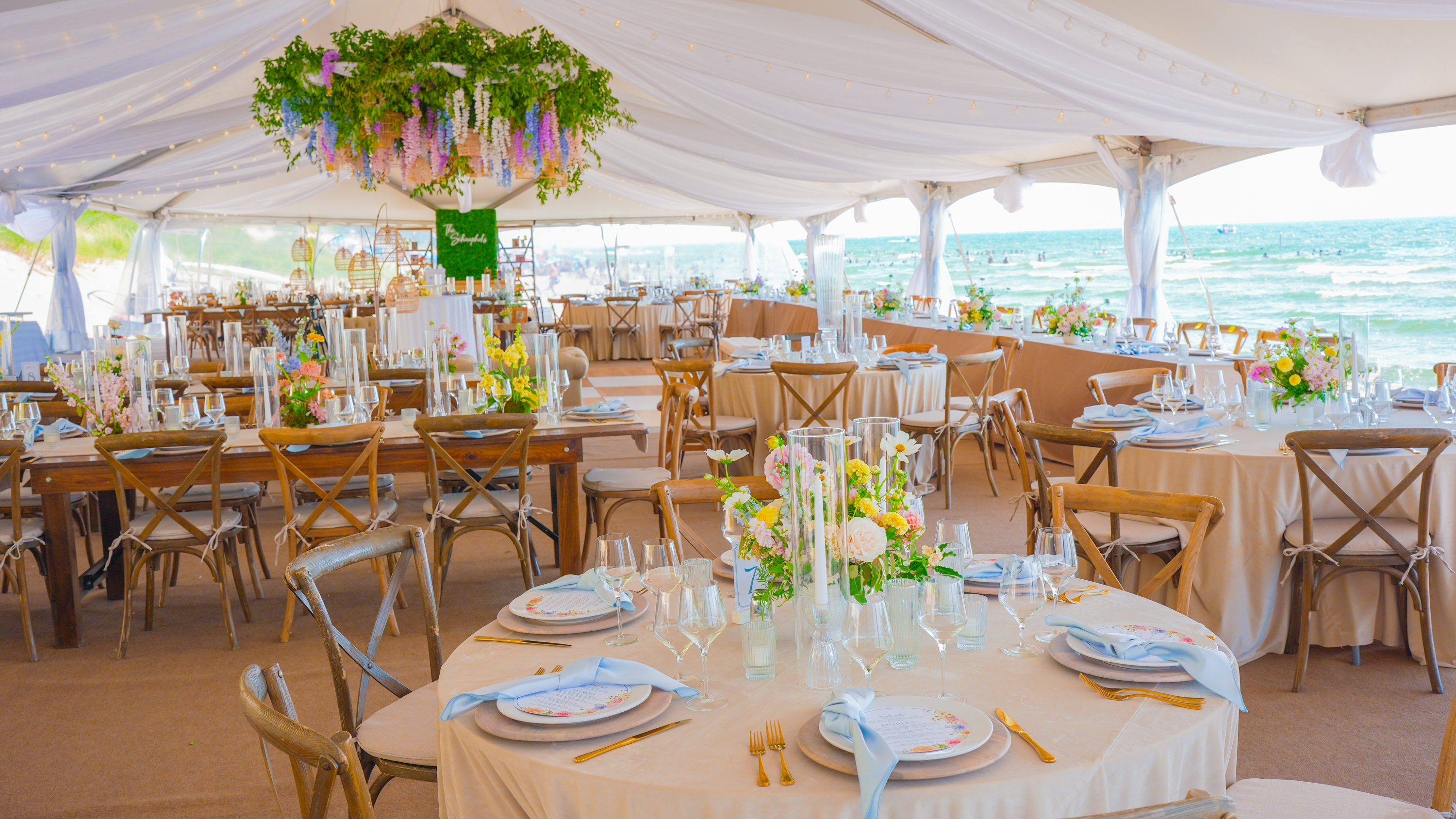 Designer-level beach wedding tent with hanging greenery chandelier, farm tables, and ocean view